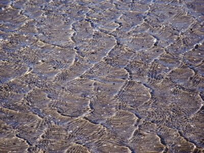 Shallow tidal water ripples over sandy beach creating intricate light-reflecting textures in a coastal close-up photograph