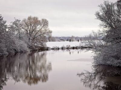 Snowy riverside winter landscape with leafless trees reflected in calm water and two people walking on a distant bank