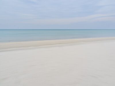Soft minimalist seascape photograph of a pale sandy beach meeting calm turquoise sea under a cloudy blue sky