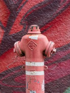 Close-up of weathered red fire hydrant with white bands and graffiti-covered red wall background, urban street photography
