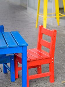 Bright red wooden child's chair beside a blue wooden table on grey pavement with a yellow stool blurred in background