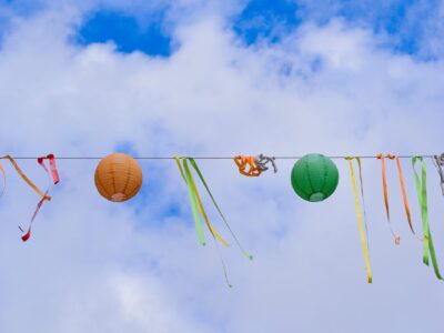 Colorful orange and green paper lanterns with long ribbon streamers hung on a wire against a bright cloudy sky