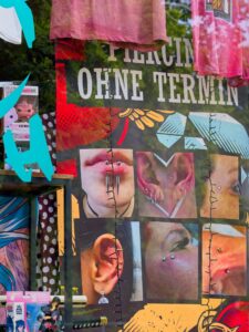 Colourful piercing shop window showing close-up photos of ear, lip and facial piercings with hanging T‑shirts and reflections