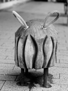 Black-and-white close-up of a carved wooden bollard with two looped ropes through metal eyes in Delmenhorst