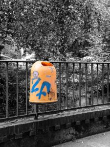 Yellow graffiti-covered street bin with blue spray paint mounted on black metal railing in leafy Delmenhorst park scene