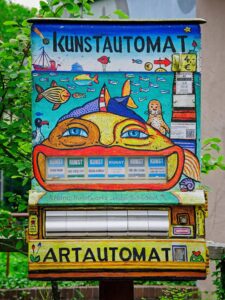 Colourful street art vending box labelled KUNSTAUTOMAT and ARTAUTOMAT with painted fish, big face and coin slot, Delmenhorst