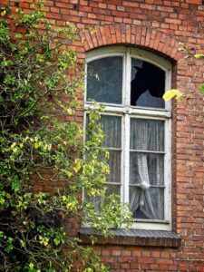 Weathered white arched sash window with a broken top pane, lace curtains and ivy climbing a red brick Delmenhorst house