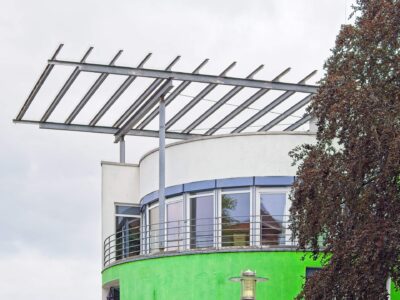 Modern Delmenhorst building with curved balcony, green facade, metal pergola roof structure and leafy tree against grey sky