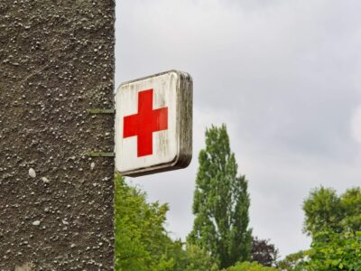 Weathered illuminated Red Cross clinic sign mounted on a textured concrete wall with trees and cloudy sky background
