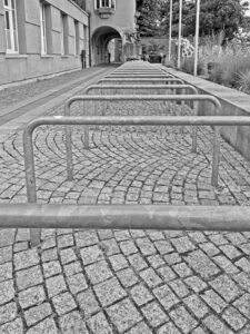 Empty bike racks on circular cobblestone pavement beside a historic municipal building and landscaped garden, black-and-white